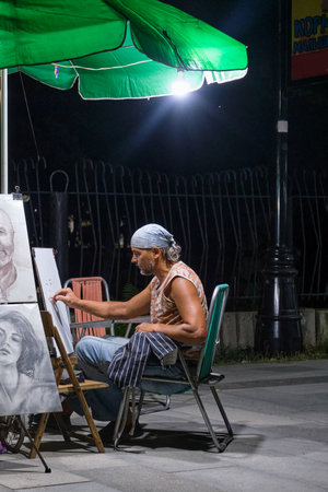 Sudak, Russia - September 15, 2020: An artist of mature age in a headscarf draws portraits on a city street in the evening under a lantern.のeditorial素材