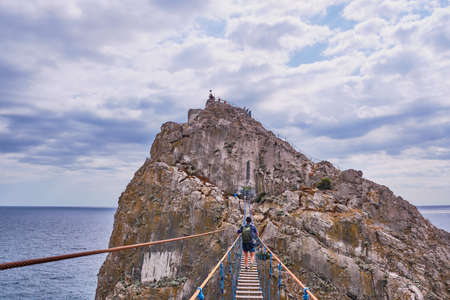 A middle-aged man with a backpack walks over a suspension bridge to Diva Rock, Black Sea coast, near Yalta, Crimea.の写真素材