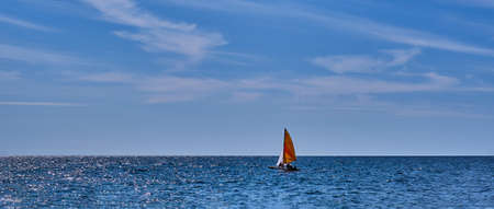 A boat with an orange-red sail floats near the rocky coast over the backdrop of the blue sea and cloudy sky.の写真素材