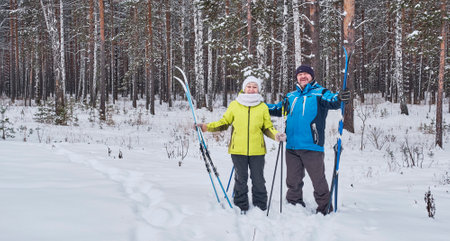 Happy mature couple in winter sportswear standing with cross-country skis in snowy forest and smilingの写真素材