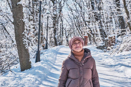 Portrait of a senior woman in a brown jacket, pink fur cap walking and looking up at something against the background of a winter snowy parkの写真素材