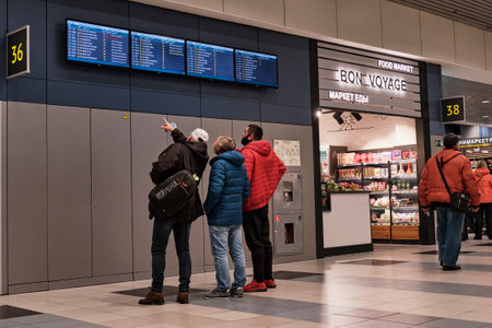 In the airport building, three male passengers in jackets and protective masks stand and watch attentively on the arrival and departure board.のeditorial素材