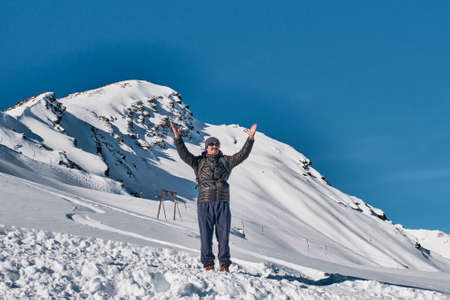 Winter mountain landscape. A mature man with a backpack in warm sports clothes and sunglasses raised his hands in greeting against the background of the top of Cheget mountain, North Caucasus, Russia.の写真素材