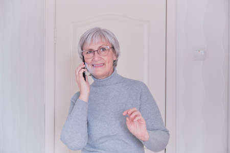 A gray-haired senior woman with glasses and a gray turtleneck talking on mobile phone at home. People connection, communication. Lifestyle moment.の写真素材