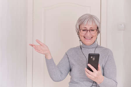 A gray-haired senior woman with glasses and a gray turtleneck talking on mobile phone at home. People connection, communication. Lifestyle moment.の写真素材