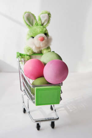 Toy soft green rabbit and a small shopping cart with colored natural chicken eggs.の写真素材