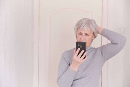 A gray-haired senior woman in a gray turtleneck looking attentively at the smartphone at home. Lifestyle moment.の写真素材