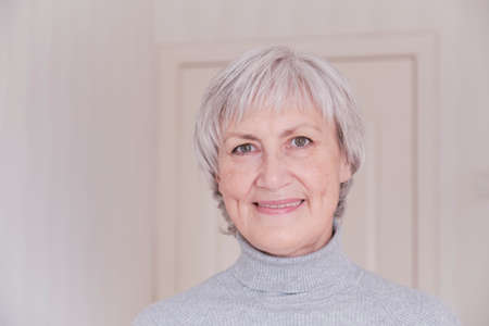 A close-up portrait of a smiling and looking at the camera elderly Caucasian woman with short gray hair on a light background.の写真素材