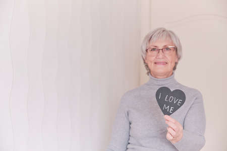 A portrait of an elderly Caucasian woman with short gray hair wearing glasses holding in her hand a black heart with the inscription I love me. Selective focus on the heart.の写真素材