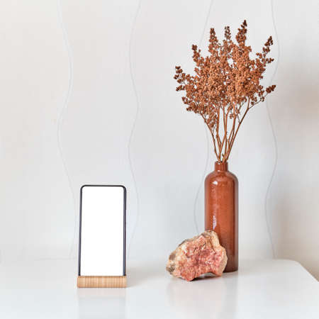 A smartphone, natural stone, a clay vase with brown dry flowers on a white coffee table against a white wall.の写真素材