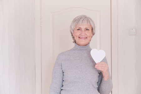 A portrait of a smiling elderly Caucasian women with short gray hair holding in your hand a small white heart.の写真素材