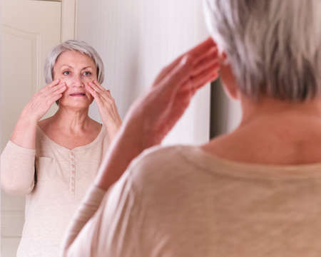A elderly Caucasian women with short gray hair in casual clothes cares for the skin of the face, makes self-massage.の写真素材