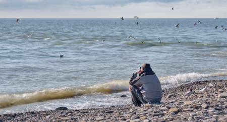 A senior man in a jacket and knitted hat sits on a rocky beach near the cold seaの写真素材