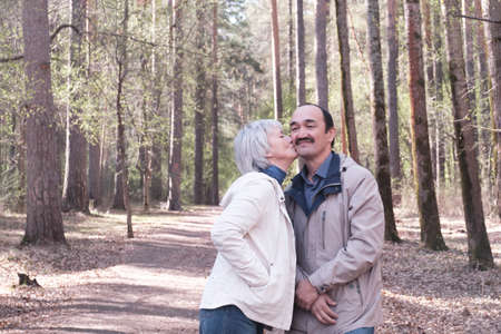 Elderly interracial couple in a spring forest park. A woman kissing a man on the cheek.の写真素材