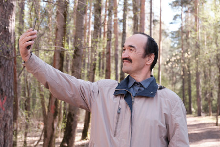 Senior Asian man using mobile phone for taking selfies in forest park.の写真素材