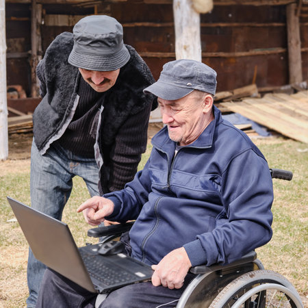 Happy senior man in a wheelchair in the yard of his house and his friend smiling, looking at a laptop.の写真素材
