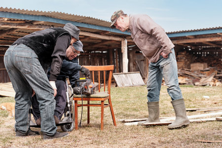 Elderly man in a wheelchair is repairing an old electric saw in his yard. His two friends are watching attentively.の写真素材