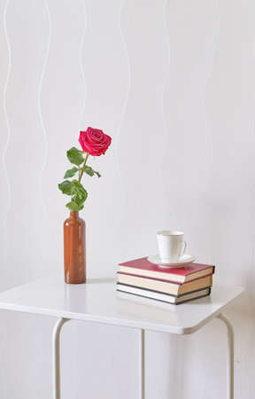 Gentle pink rose in clay bottle, cup of coffee, books on a white coffee table against a light wall.の写真素材
