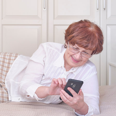 Senior woman wearing glasses, using mobile phone on the bed at home.の写真素材
