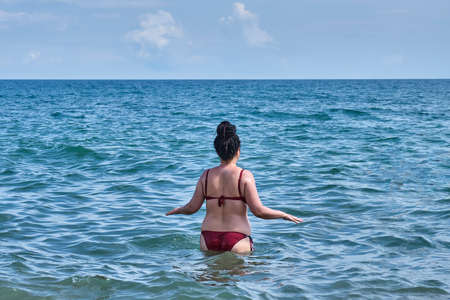 A young asian woman in a red swimsuit walking into the sea. Summer seascape.の写真素材