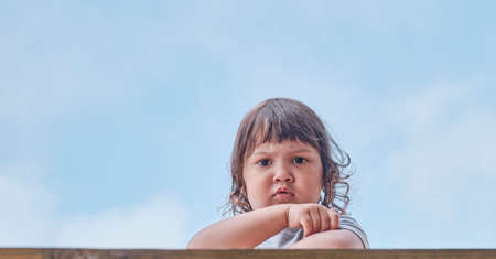 Portrait of cute little Asian frowning baby girl with dark hair on blue sky background.の写真素材