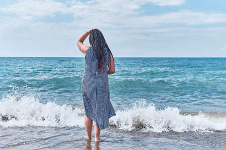 A young woman with dreadlocks on black hair, in baseball cap and long striped dress walking into the water.の写真素材