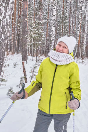 Happy smiling senior woman in yellow jacket, white knitted hat and scarf with ski poles over background of winter snowy forestの写真素材