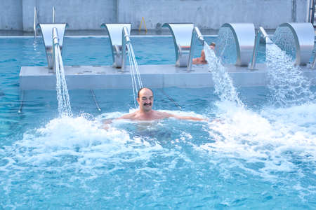 Happy mature asian man doing massage with water jets from injector in outdoor thermal pool.