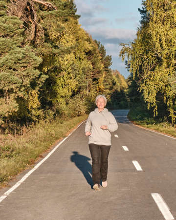 Senior active woman with gray hair running along forest paved road on sunny autumn day.の写真素材