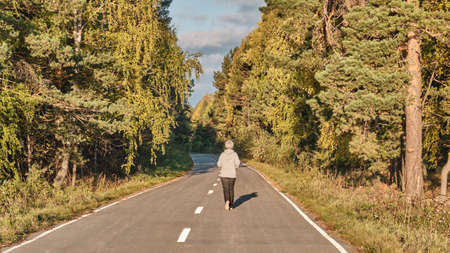 Senior active woman with gray hair running along forest paved road on sunny autumn day.の写真素材