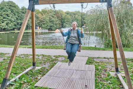 Happy senior caucasian woman with short gray hair swinging on chain swing in parkの写真素材