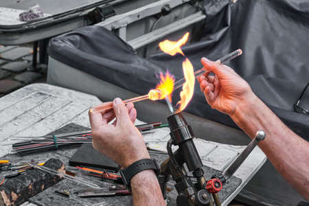 Hands of mature glassblower holding glass rod over burner flame. Process of artistic glass processing, flamework.の写真素材