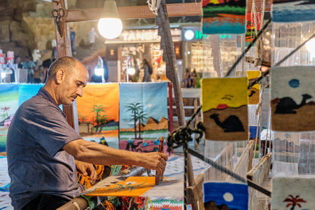 Middle aged man on night street spinning colorful small rug as tourist souvenir on handloom.のeditorial素材