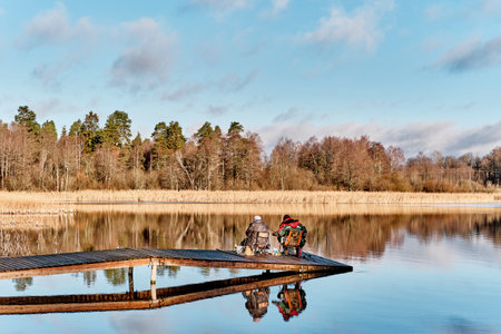 Two fisherman with fishing rods sitting on wooden bridge among scenic lake, reflecting on mirror surface of water.の写真素材