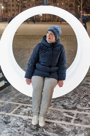 Portrait of senior woman in ice skates resting on ring bench at city skating rink on winter evening.の写真素材