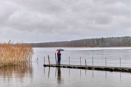 Man standing under umbrella on wooden pier on autumn rainy gray day over of backdrop of lake and forest.の写真素材