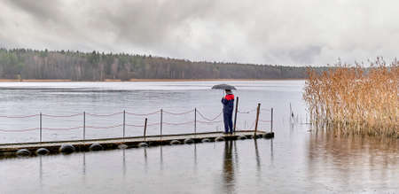 Man standing under umbrella on wooden pier on autumn rainy gray day over of backdrop of lake and forest.の写真素材