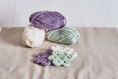 Colorful flowers knitted of crochet. Skeins of yarn, light green, violet and white on linen napkin.