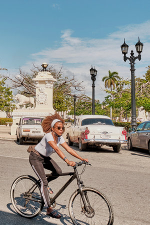 Young cute black woman in sportswear and sunglasses riding bicycle in street of Cienfuegos, Cubaのeditorial素材