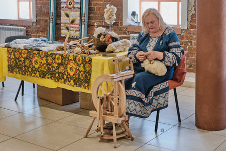 Senior Caucasian woman in traditional Russian dress spinning on wooden spinning wheel.のeditorial素材