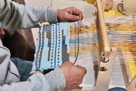 Hands of senior man weaving small rug with pattern on manual table loom.