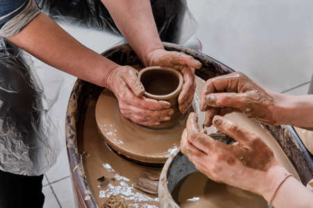 Hands of senior woman and girl sculpting clay vase on potters wheel at pottery training lesson.