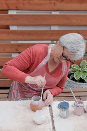 Senior gray haired woman with glasses covering with white glaze handmade clay vase by brushの写真素材
