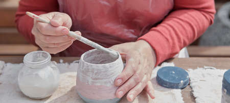 Hands of older woman painting handmade clay vase with colored paints with brush.