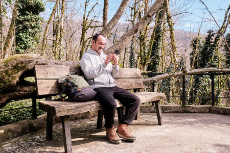 Senior asian man with backpack uses a cell phone while sitting on wooden bench at resting place in relict forest. Yew-boxwood grove, Caucasian Reserve, Russia.の写真素材