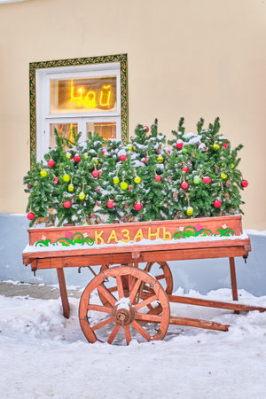 Outdoor decoration for winter holidays. On bright painted wooden old cart are spruce branches with Christmas balls. Inscription in Russian - Kazan. Snow-covered street of city of Kazan, Russiaの写真素材