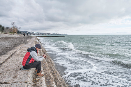 Senior Asian man using mobile phone while sitting on stone steps near sea on cloudy spring day. Black Sea, Sochi, Russia. technology, real people conceptの写真素材