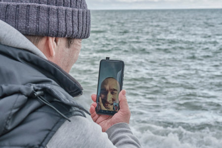 Over shoulder view of senior asian man looking at cellphone screen,talking on video call with his...