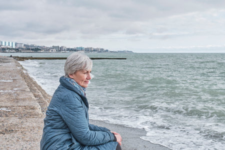 Senior woman sitting on sea coast. Retirement vacation on cold cloudy day. side view. Elderly people concept.の写真素材