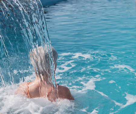 Senior woman with gray hair enjoying flow of water falling on her shoulders. rear view. Hydromassage in outdoor thermal swimming pool. Active elderly concept.の写真素材